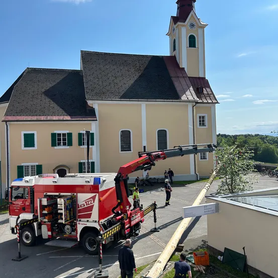 Ein roter Feuerwehrwagen mit ausgefahrener Leiter steht vor einer Kirche. Eine Person steht in der Nähe der Leiter. Ein Schild mit deutschem Text ist an einem Pfosten montiert.