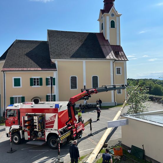 Ein roter Feuerwehrwagen mit ausgefahrener Leiter steht vor einer Kirche. Eine Person steht in der Nähe der Leiter. Ein Schild mit deutschem Text ist an einem Pfosten montiert.