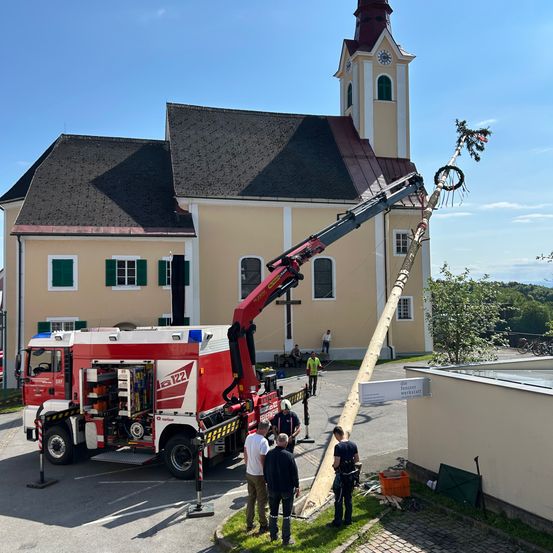 Ein Kran steht vor einer Kirche und hebt einen hohen Holzpfahl. Mehrere Personen beobachten, einige in Sicherheitswesten. Ein roter Feuerwehrwagen ist in der Nähe geparkt.