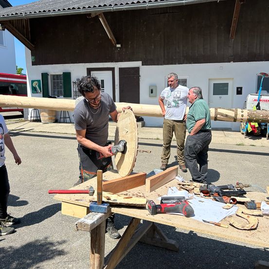 Ein Mann mit lockigen Haaren benutzt ein Elektrowerkzeug, um ein großes Stück Holz auf einem Werktisch zu schnitzen. Hinter ihm beobachten und unterhalten sich zwei Männer.