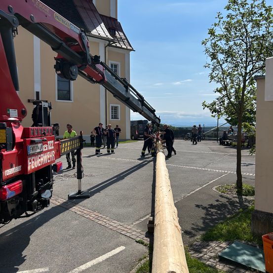 Ein Kran wird benutzt, um einen langen Holzpfahl in einem Parkplatz zu heben. Mehrere Menschen sind in Helmen und Feuerwehruniformen versammelt. Ein Gebäude mit Fenstern und ein Baum sind im Hintergrund zu sehen. Der Himmel ist blau mit einigen Wolken.