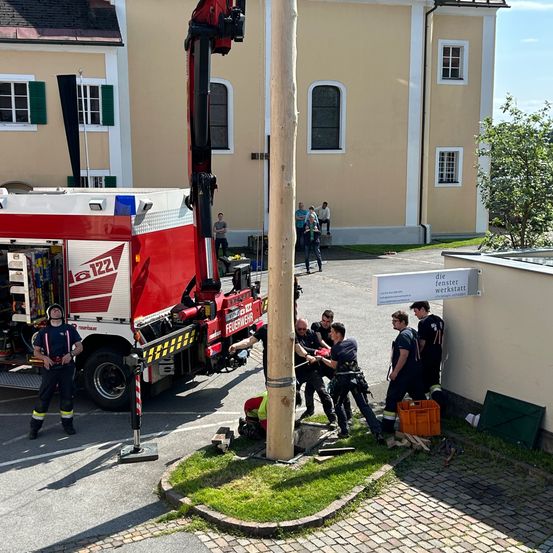 Eine Gruppe Feuerwehrleute arbeitet an einem Pfahl mit einem Kran dahinter. Sie stehen auf dem Ziegelpflaster in der Nähe eines Gebäudes. Ein Feuerwehrwagen ist nebenan geparkt.
