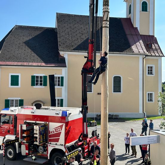 Ein Mann klettert an einem hohen Holzpfahl, der mit Seilen gesichert ist. Er steht auf einem roten Kran neben einem Feuerwehrfahrzeug mit offener Hecktür. Mehrere Menschen stehen herum und beobachten. Im Hintergrund befindet sich ein Gebäude mit grünen Fensterläden.