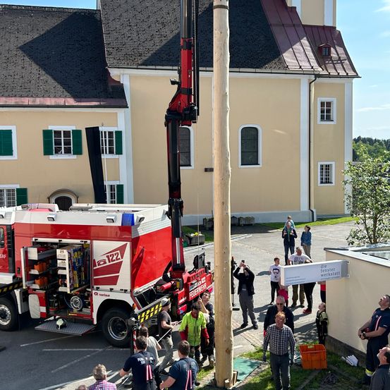Ein roter Kran wird verwendet, um einen Holzpfahl vor einer Kirche anzuheben. Mehrere Personen beobachten den Kran und den Pfahl.
