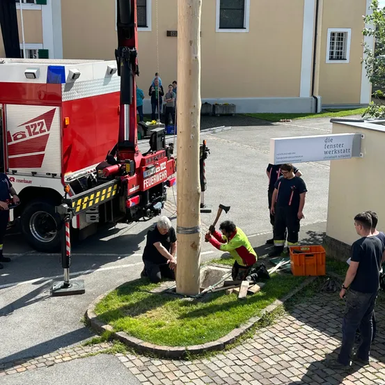 Eine Gruppe von Feuerwehrleuten arbeitet auf einer Straße und benutzt einen großen roten Lastwagen und einen Pfosten. Ein Mann schlägt auf den Pfosten, während andere zuschauen. Ein Gebäude mit einem Schild, das 'die fenster werkstatt' besagt, befindet sich in der Nähe.