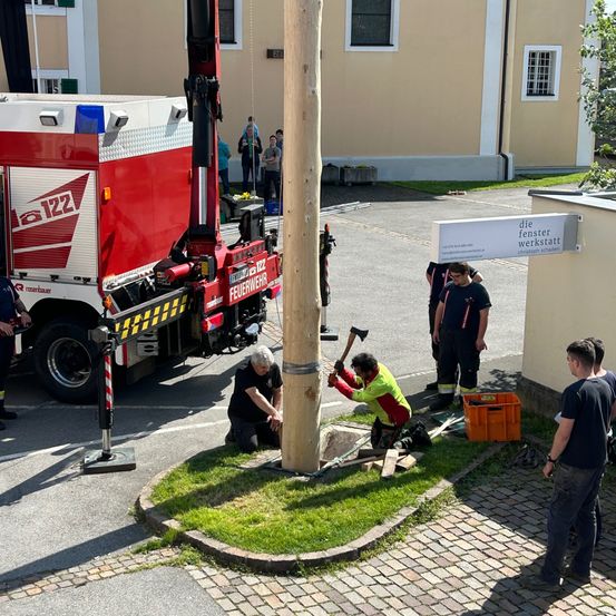 Eine Gruppe von Feuerwehrleuten arbeitet auf einer Straße und benutzt einen großen roten Lastwagen und einen Pfosten. Ein Mann schlägt auf den Pfosten, während andere zuschauen. Ein Gebäude mit einem Schild, das 'die fenster werkstatt' besagt, befindet sich in der Nähe.