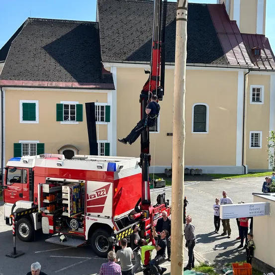 Ein Feuerwehrmann benutzt einen Kran, um einen hohen Mast vor einem Gebäude mit rotem Dach zu erklimmen.