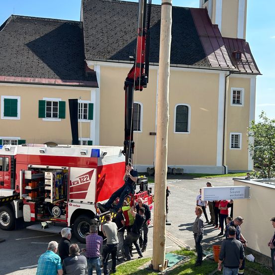 Ein Feuerwehrmann benutzt einen Kran, um die Spitze eines hohen Pfahls vor einem Gebäude zu erreichen.