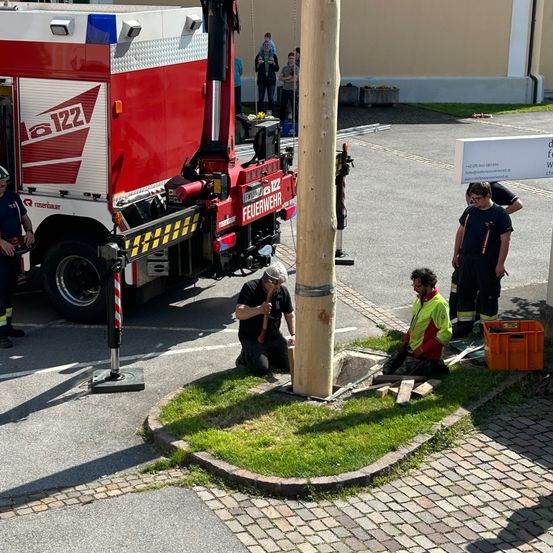 Eine Gruppe von Feuerwehrleuten arbeitet auf einer Straße und repariert einen Hydranten. Ein Mann schlägt mit einem Hammer auf eine Rohrleitung. Ein anderer Mann sitzt auf dem Rasen. Ein Feuerwehrwagen ist auf der Straße geparkt, mit Menschen, die dahinter stehen.