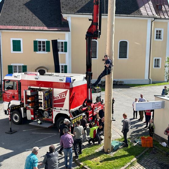 Ein Feuerwehrmann klettert mithilfe eines Krans einen hohen Pfahl. In der Nähe ist ein Feuerwehrwagen geparkt, mit offenem Heck, das Ausrüstung im Inneren zeigt. Menschen sind versammelt, einige tragen Westen und andere stehen in der Nähe eines Gebäudes mit mehreren Fenstern.