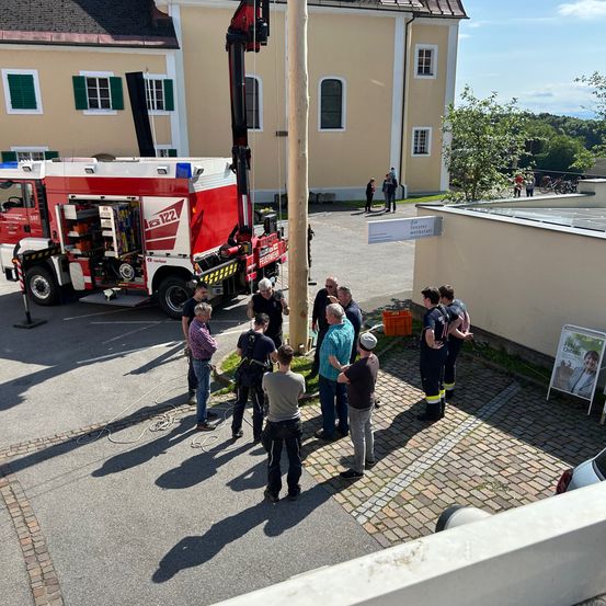 Eine Gruppe von Menschen steht vor einem roten Feuerwehrwagen mit offener Tür und angebrachtem Kran. Ein Mann steht in der Nähe des Krans. Die Szene spielt vor einem Gebäude mit gelber Wand, einem Baum und einem Schild.
