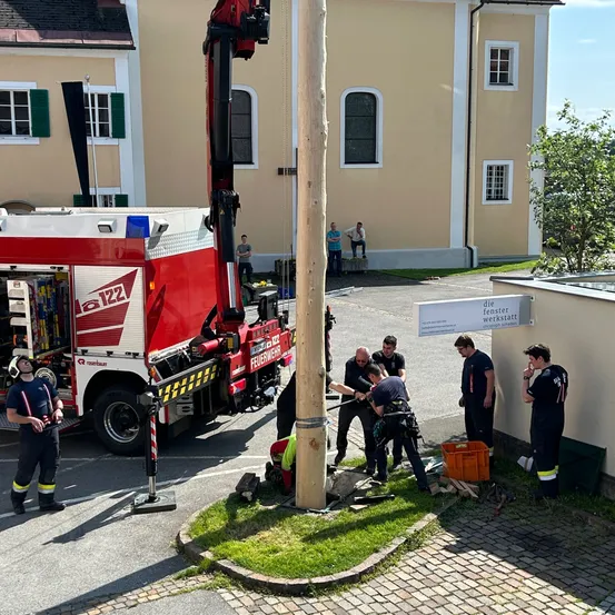 Eine Gruppe von Menschen arbeitet an einem Holzpfahl vor einem Gebäude. Ein Feuerwehrwagen ist auf der linken Seite geparkt, und ein Aufbewahrungsbox steht auf dem Boden. Einige Leute stehen im hinteren Teil des Gebäudes.