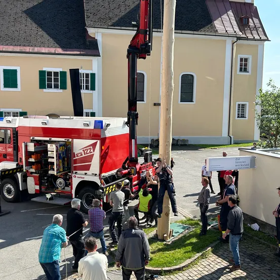 Eine Gruppe von Menschen steht um einen Feuerwehrwagen herum, der vor einem Gebäude mit dem Schild 'Die Fensterwerkstatt' geparkt ist. Der Feuerwehrwagen steht am Straßenrand.