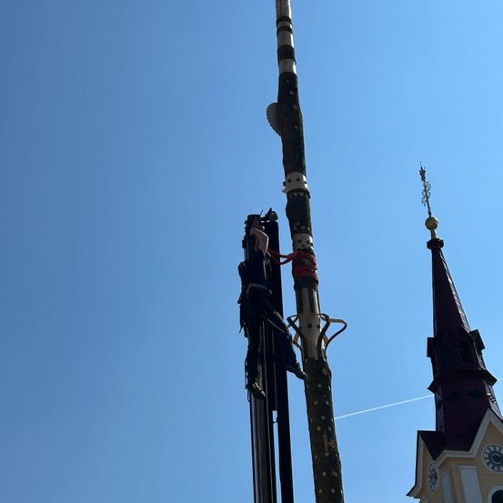 Eine Person klettert an einem sonnigen Tag vor einer Kirche an einem hohen Holzpfahl hoch.
