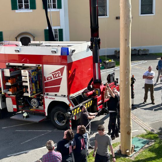 Ein roter und weißer Feuerwehrwagen ist am Straßenrand geparkt, wobei Feuerwehrleute einen Kran benutzen, um einen hohen Holzpfahl anzuheben. Mehrere Menschen beobachten die Aktivität. Die Feuerwehrleute tragen Helme und Uniformen, und im Hintergrund befindet sich ein Gebäude mit grünen Fensterläden.