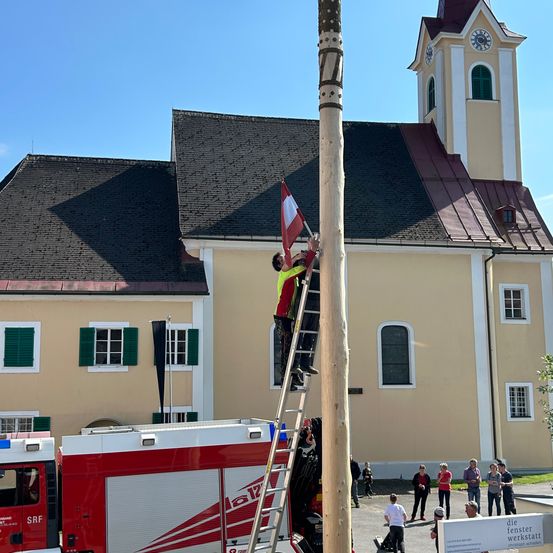 Eine Person klettert auf eine Leiter, um eine Flagge vor einem Gebäude mit Turm und Uhr zu hissen.