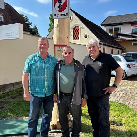 Drei Männer stehen vor einem Holzpfahl mit einem Schild, das 'FF ST.NIKOLAI' besagt, lächelnd, mit Häusern und geparkten Autos im Hintergrund.