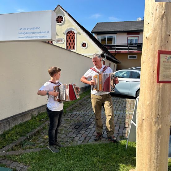 Ein Mann und ein Junge spielen Akkordeons in einem Innenhof, mit einem geparkten Auto und einem Gebäude im Hintergrund.