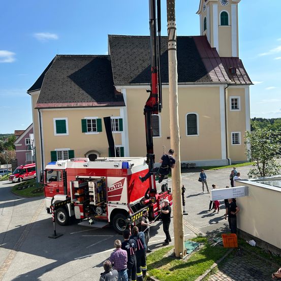 Eine Gruppe von Menschen steht vor einer Kirche. Ein Feuerwehrmann klettert mit einem Kran auf einen Pfahl. Der Feuerwehrwagen ist vor der Kirche geparkt.