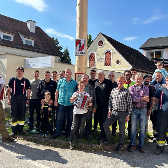 Eine Gruppe von Menschen, einschließlich Feuerwehrleuten, steht vor einem Gebäude mit einem Schild, auf dem 'FF ST.NIKOLAI' steht. Einige tragen Uniformen und Mützen, und ein Kind hält ein Akkordeon.