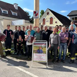 Eine Gruppe von Erwachsenen und Kindern, einige in Uniformen, stehen draußen vor einem Haus mit einem Schild und einem Plakat vor ihnen.
