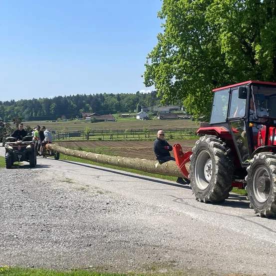 Ein Traktor zieht einen langen Baumstamm auf einer Landstraße, mit mehreren Personen, die auf einem Quad und einem Motorrad dahinter fahren. Im Hintergrund befindet sich ein weitläufiges Feld mit einem Baum und mehreren Häusern.