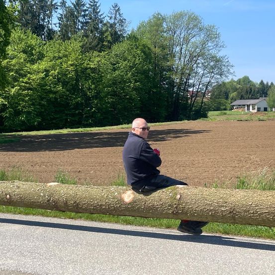 Ein Mann mit Sonnenbrille sitzt auf einem großen Baumstamm am Straßenrand.