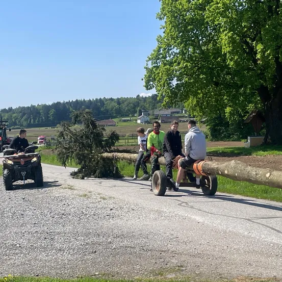 Vier junge Männer sitzen auf einem Baumstamm, der von einem Traktor gezogen wird. Der Traktor fährt auf einer Kiesstraße, vor ihm fährt ein Mann mit einem ATV.