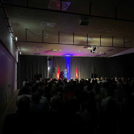 Ein Mann steht mit einem Mikrofon auf der Bühne und spricht eine große Zuhörerschaft in einem abgedunkelten Saal an. Die Bühne ist mit blauen und roten Lichtern beleuchtet.