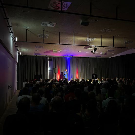 Ein Mann steht mit einem Mikrofon auf der Bühne und spricht eine große Zuhörerschaft in einem abgedunkelten Saal an. Die Bühne ist mit blauen und roten Lichtern beleuchtet.