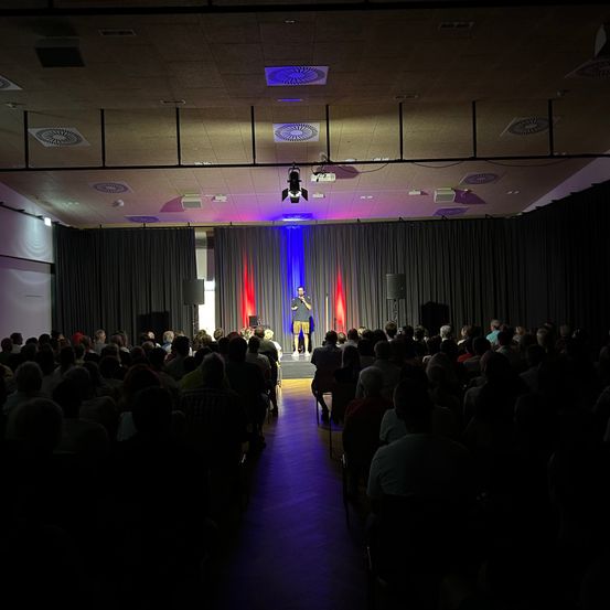 Ein Mann steht mit einem Mikrofon auf der Bühne vor einem sitzenden Publikum. Die Bühne ist mit einem rot-blauen Vorhang geschmückt, und Scheinwerfer beleuchten die Szene.