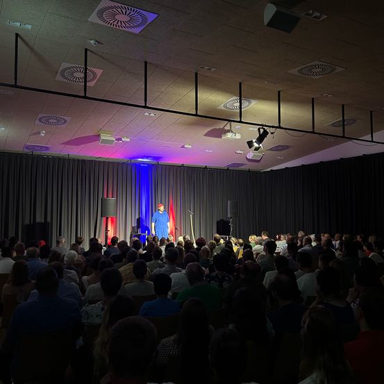 Ein Mann steht mit einem Mikrofon auf der Bühne und spricht eine große Zuhörerschaft in einem Raum mit schwarzem Vorhang und farbiger Beleuchtung an.