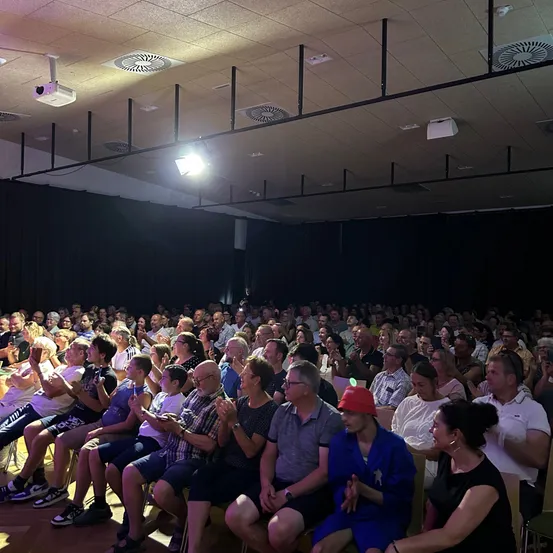 Eine große Menschenmenge sitzt in einem Auditorium, viele klatschen. Der Raum hat eine Bühne, schwarze Vorhänge und Deckenleuchten. Ein Scheinwerfer beleuchtet die Vorderseite.