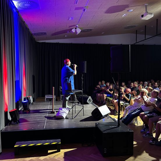 Eine Person auf der Bühne mit einem Mikrofon, die für ein Publikum auftritt, das in Stühlen und auf dem Boden sitzt. Die Bühne hat Lautsprecher, und Beleuchtungsequipment ist sichtbar.