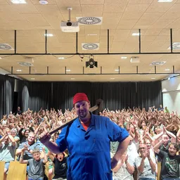 Ein Mann in einem blauen Hemd und rotem Hut hält einen großen Hammer vor einer großen Zuhörerschaft. Das Publikum jubelt und hebt die Hände. Der Raum hat einen Projektor und Beleuchtung an der Decke.
