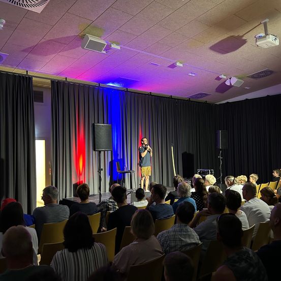 Ein Mann steht mit einem Mikrofon auf der Bühne, umgeben von bunten Lichtern. Ein Publikum sitzt in Stühlen und beobachtet ihn aufmerksam.