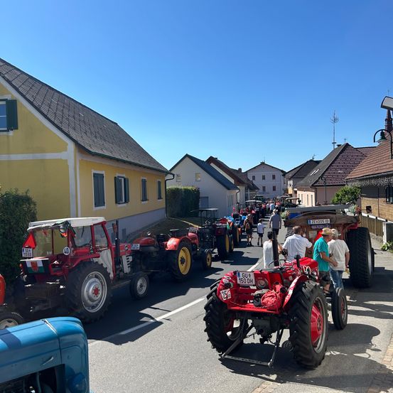 Viele rote Traktoren sind an einem sonnigen Tag in einer Reihe geparkt. Mehrere Menschen stehen in der Nähe der Traktoren, einer sitzt auf einem. In der Nähe befinden sich mehrere Häuser, eines davon hat einen Schornstein.