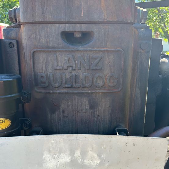Eine Nahaufnahme eines verrosteten Metallobjekts, möglicherweise ein Motorteil, mit der Aufschrift 'LANZ BULLDOG'. Es hat Schrauben und ein Loch in der Mitte.