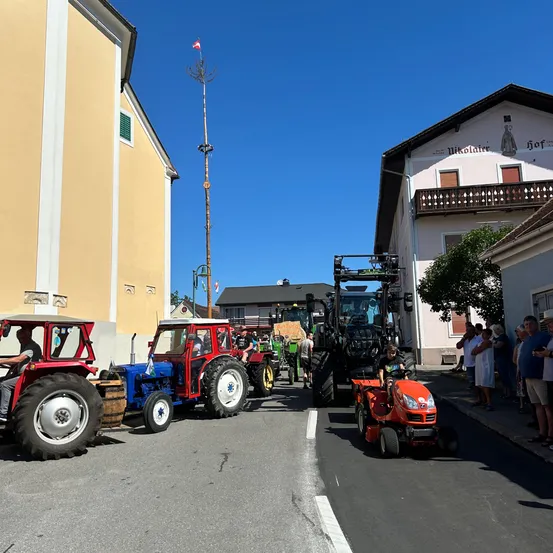 Eine Parade von Traktoren, darunter ein roter, blauer und schwarzer, fährt eine Straße in einem Dorf hinunter. Zuschauer beobachten vom Bürgersteig aus. Gebäude säumen die Straße, und ein Fahnenmast steht in der Nähe.
