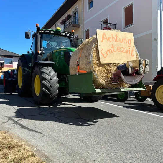 Ein grüner Traktor fährt auf der Straße mit einem Heuballen am Heck, und eine Person liegt darauf mit dem Schild 'Achtung Erntezeit'. Dahinter steht ein Gebäude mit Balkon und Fenstern. Eine Person sitzt auf dem Traktor.