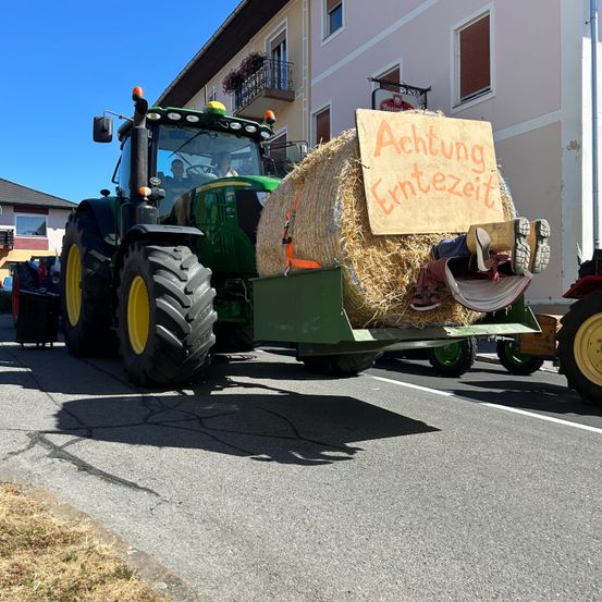 Ein grüner Traktor fährt auf der Straße mit einem Heuballen am Heck, und eine Person liegt darauf mit dem Schild 'Achtung Erntezeit'. Dahinter steht ein Gebäude mit Balkon und Fenstern. Eine Person sitzt auf dem Traktor.