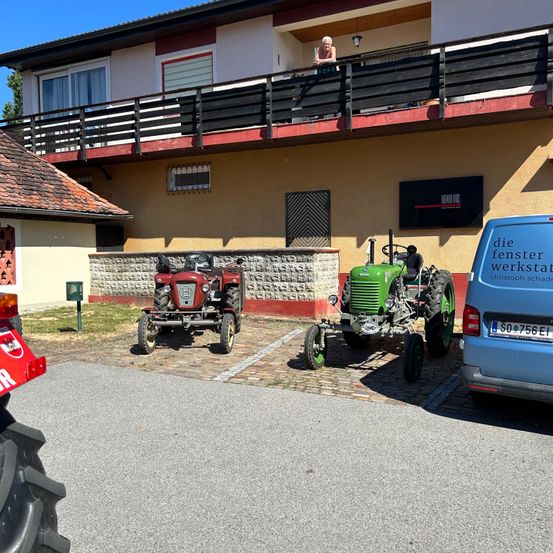 Eine Frau steht auf dem Balkon eines Gebäudes mit zwei Traktoren davor. Ein Van mit einem Nummernschild ist in der Nähe geparkt.