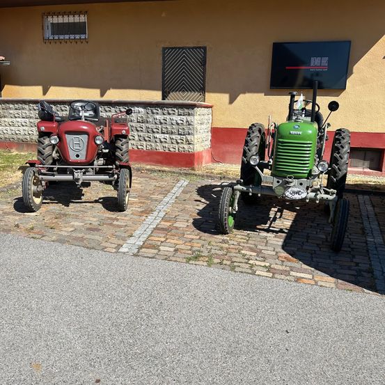Zwei Oldtimer-Traktoren, einer rot und einer grün, geparkt auf einer Ziegelstraße vor einem Gebäude mit einem Schild, das 'Oldtimer Farms' steht.