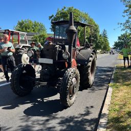 Ein alter Traktor steht am Straßenrand. Leute stehen herum, einer hält ein Tablet. Hinter dem Traktor sind weitere Menschen und ein weiterer Traktor. Im Hintergrund befinden sich Bäume und Häuser.