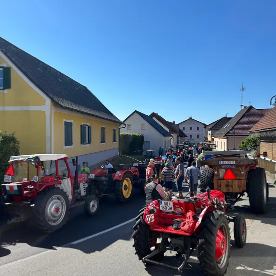 Eine Reihe roter Traktoren auf einer Straße, mit Menschen, die herumlaufen und sitzen. Die Traktoren sind aufgereiht, und einige sind geparkt. Die Gebäude in der Gegend sind gelb und braun, einige mit Fenstern. Rechts befindet sich ein Zaun.