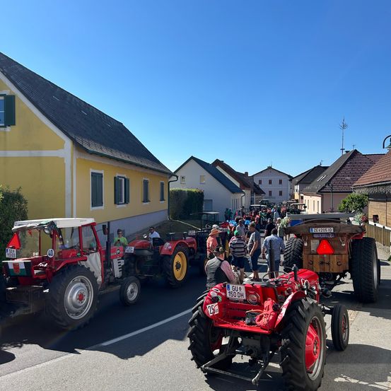 Eine Reihe roter Traktoren auf einer Straße, mit Menschen, die herumlaufen und sitzen. Die Traktoren sind aufgereiht, und einige sind geparkt. Die Gebäude in der Gegend sind gelb und braun, einige mit Fenstern. Rechts befindet sich ein Zaun.