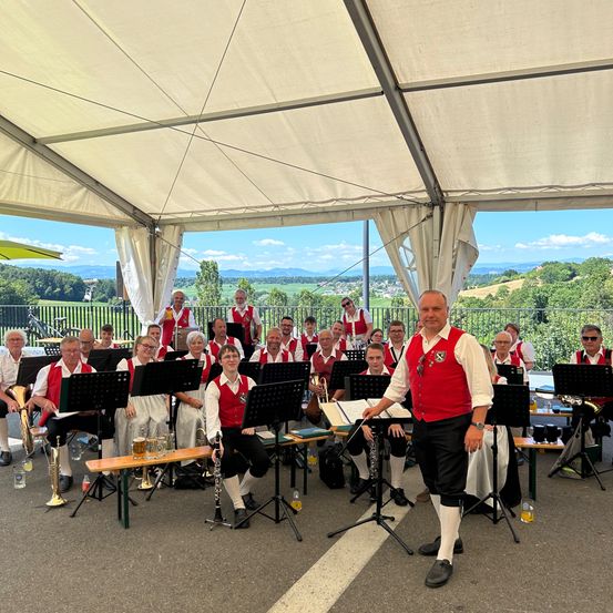 Ein Blasorchester in roten Westen und weißen Hemden spielt unter einem weißen Zelt mit malerischer Kulisse. Die Band steht in einem Halbkreis, mit einer Person vorne.