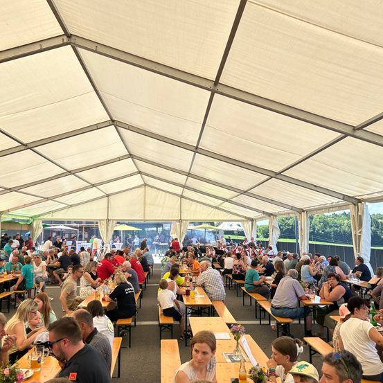 Eine große Gruppe von Menschen sitzt unter einem weißen Zelt. Viele Tische und Bänke sind mit essen und trinkenden Menschen gefüllt. Einige Personen stehen in der Nähe des Zelteingangs. Das umliegende Gebiet ist mit Bäumen gefüllt.