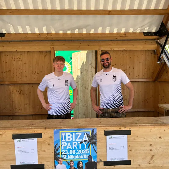 Zwei Männer in weißen Trikots mit den Nummern 3 und 11 stehen in einem Holzhäuschen und lächeln. Hinter ihnen ist ein grüner Banner und ein Fenster zu sehen. Poster für eine Ibiza-Party sind auf dem Tresen.