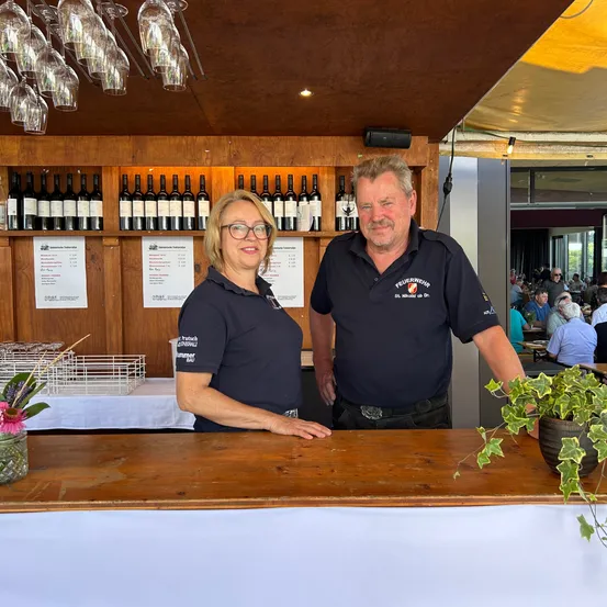 Eine lächelnde Frau und ein Mann, beide in blauen Uniformen, stehen hinter einer Bartheke in einem Restaurant. Hinter ihnen befinden sich Regale mit Weinflaschen und im Hintergrund sitzende Gäste an Tischen.
